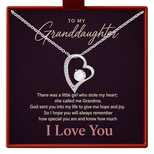 A silver heart-shaped pendant necklace with a clear center gemstone, presented on a dark burgundy gift box with sentimental text addressed to a granddaughter.