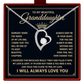 A silver heart-shaped pendant necklace with a clear central gemstone and a small crown on the chain, presented on a black gift card with gold text addressed to a granddaughter. The card is mounted inside a red gift box.