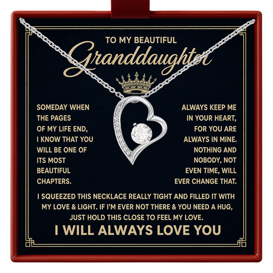 A silver heart-shaped pendant necklace with a clear central gemstone and a small crown on the chain, presented on a black gift card with gold text addressed to a granddaughter. The card is mounted inside a red gift box.