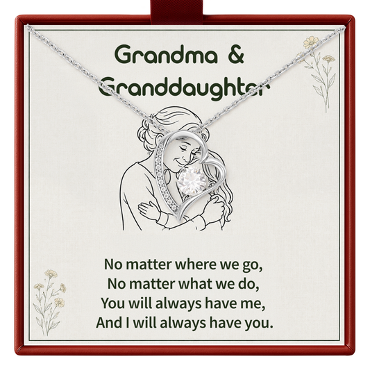 A silver heart-shaped pendant necklace with a clear gemstone, presented on a cream-colored card inside a red gift box. The card is personalized with the text 'Grandma & Granddaughter' and a sentimental quote, featuring a line drawing of the two of hugging.