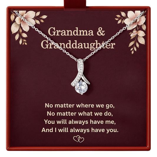Silver necklace with a curved pendant set with small clear stones and a central clear gem, displayed on a dark red gift box with floral corner details. The box is printed with the words 'Grandma & Granddaughter' and a sentimental quote.
